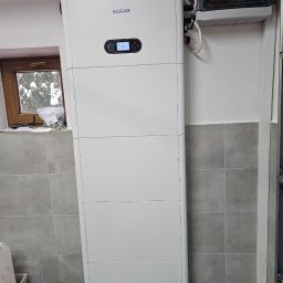 MK SUN Mateusz Karbowiak - Biały, pionowy magazyn energii SOFAR zamontowany w pomieszczeniu z szarą glazurą. Widoczna skrzynka elektryczna i okno z zimowym krajobrazem.