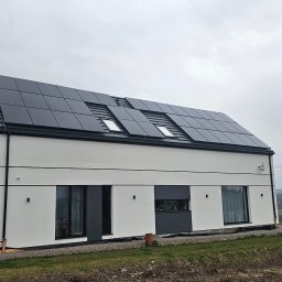 MK SUN Mateusz Karbowiak - Nowoczesny dom jednorodzinny z panelami fotowoltaicznymi na dachu, widok z zewnątrz w pochmurny dzień. Minimalistyczna architektura, ciemne ramy okienne.