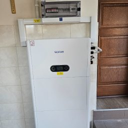 MK SUN Mateusz Karbowiak - Biały inwerter Sofar zamontowany w pomieszczeniu z brązowymi drzwiami i rozdzielnicą elektryczną. Widoczne kable i elementy instalacji.