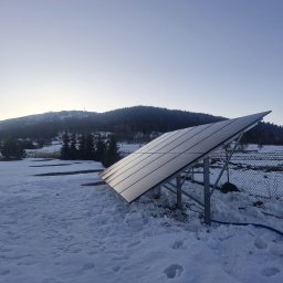 MK SUN Mateusz Karbowiak - Panel fotowoltaiczny na metalowej konstrukcji, pokryty śniegiem, z widokiem na zalesione wzgórze w tle. Poranek, delikatne światło.