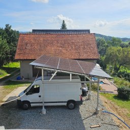 MK SUN Mateusz Karbowiak - Instalacja fotowoltaiczna nad samochodem dostawczym na tle domu z czerwoną dachówką i zieleni. Nietypowe rozwiązanie z panelami słonecznymi.