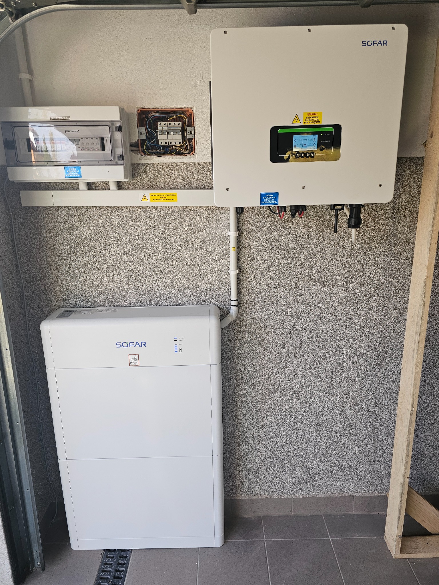 Kompletna instalacja z białym inwerterem i magazynem energii firmy SOFAR, zamontowana na szarej ścianie z widoczną skrzynką elektryczną i zabezpieczeniami, rurami PCV i drewnianą konstrukcją w rogu