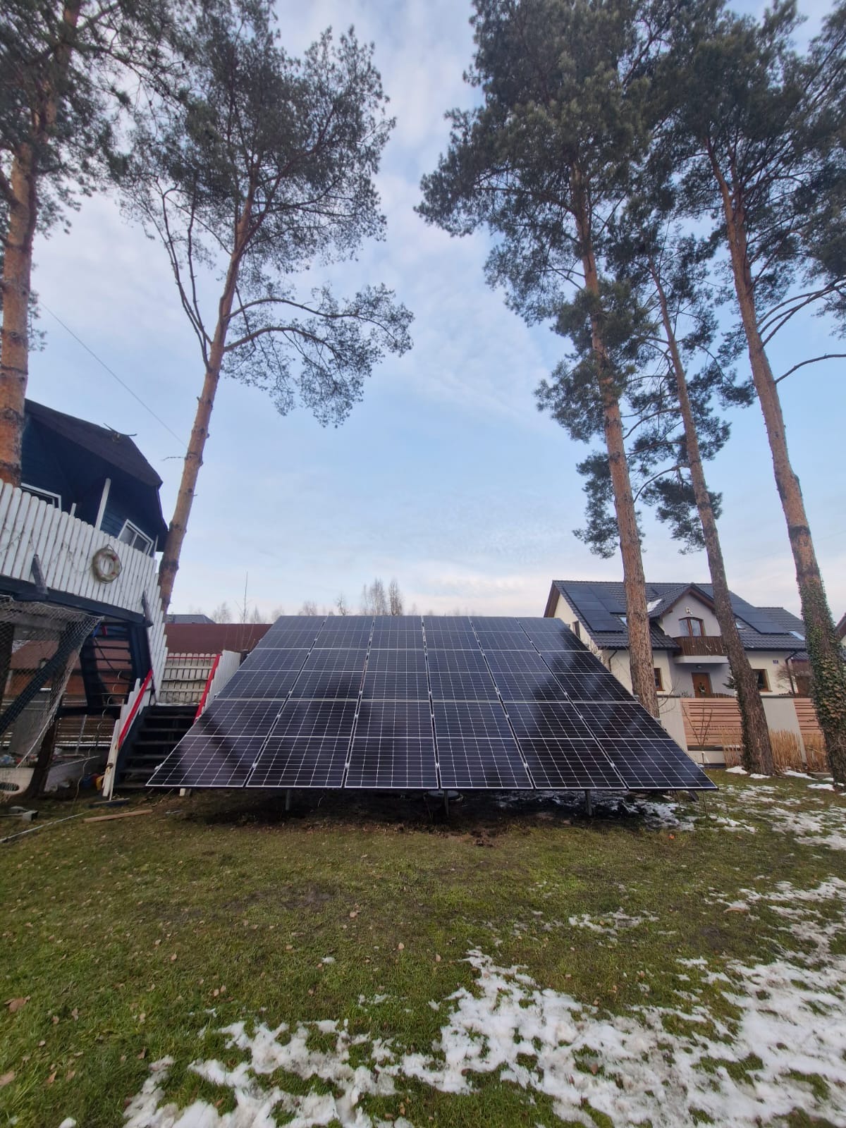 Panel słoneczny ustawiony na trawniku przed domem, otoczony wysokimi sosnami i z widocznymi panelami na dachu sąsiedniego budynku.