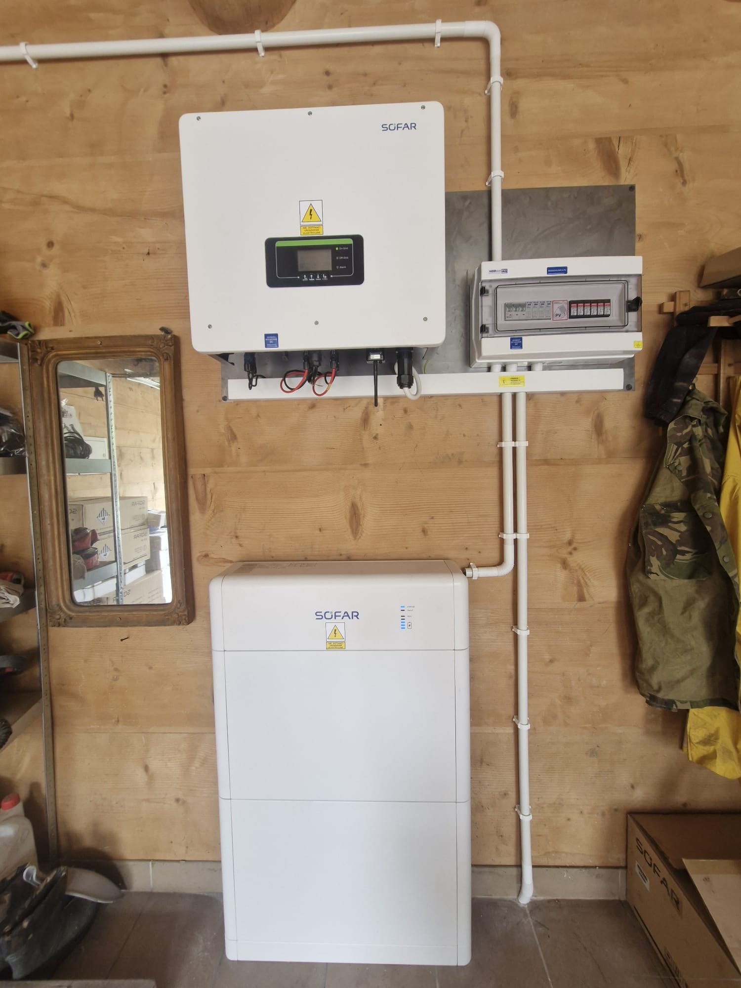 Instalacja fotowoltaiczna Sofar na drewnianej ścianie: inwerter, bateria i skrzynka z bezpiecznikami. Widoczne rury instalacyjne i lustro w ozdobnej ramie.