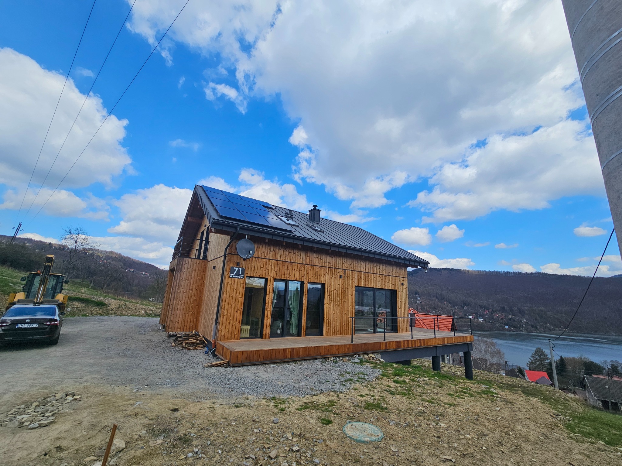 Nowoczesny, drewniany dom z panelami słonecznymi na dachu, z tarasem i widokiem na jezioro, w słoneczny dzień. Na podjeździe stoi samochód i koparka.