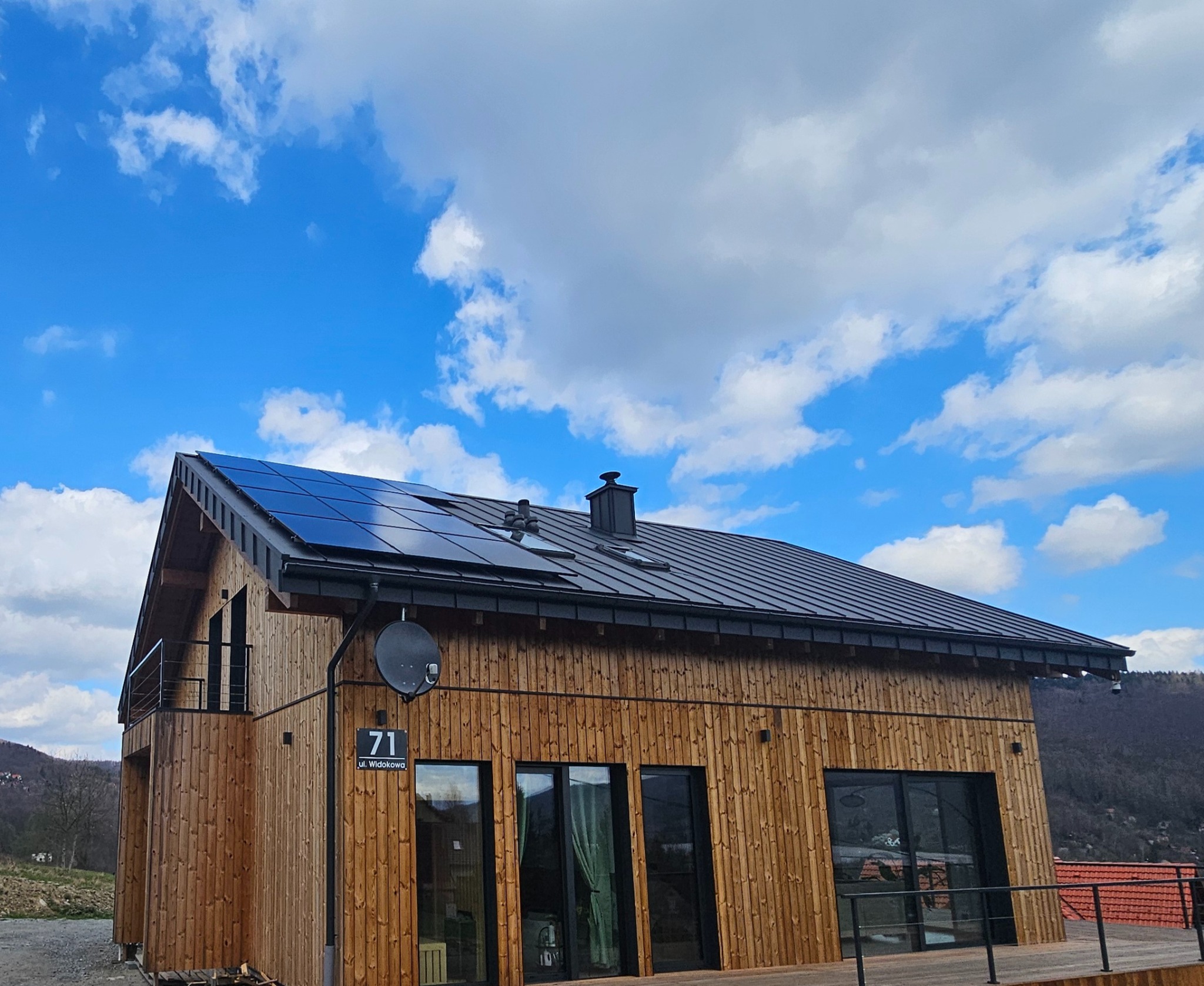 Drewniany dom z panelami słonecznymi na dachu, widok z dołu na tle błękitnego nieba z chmurami. Widoczny numer domu '71 ul. Widokowa' i satelita.