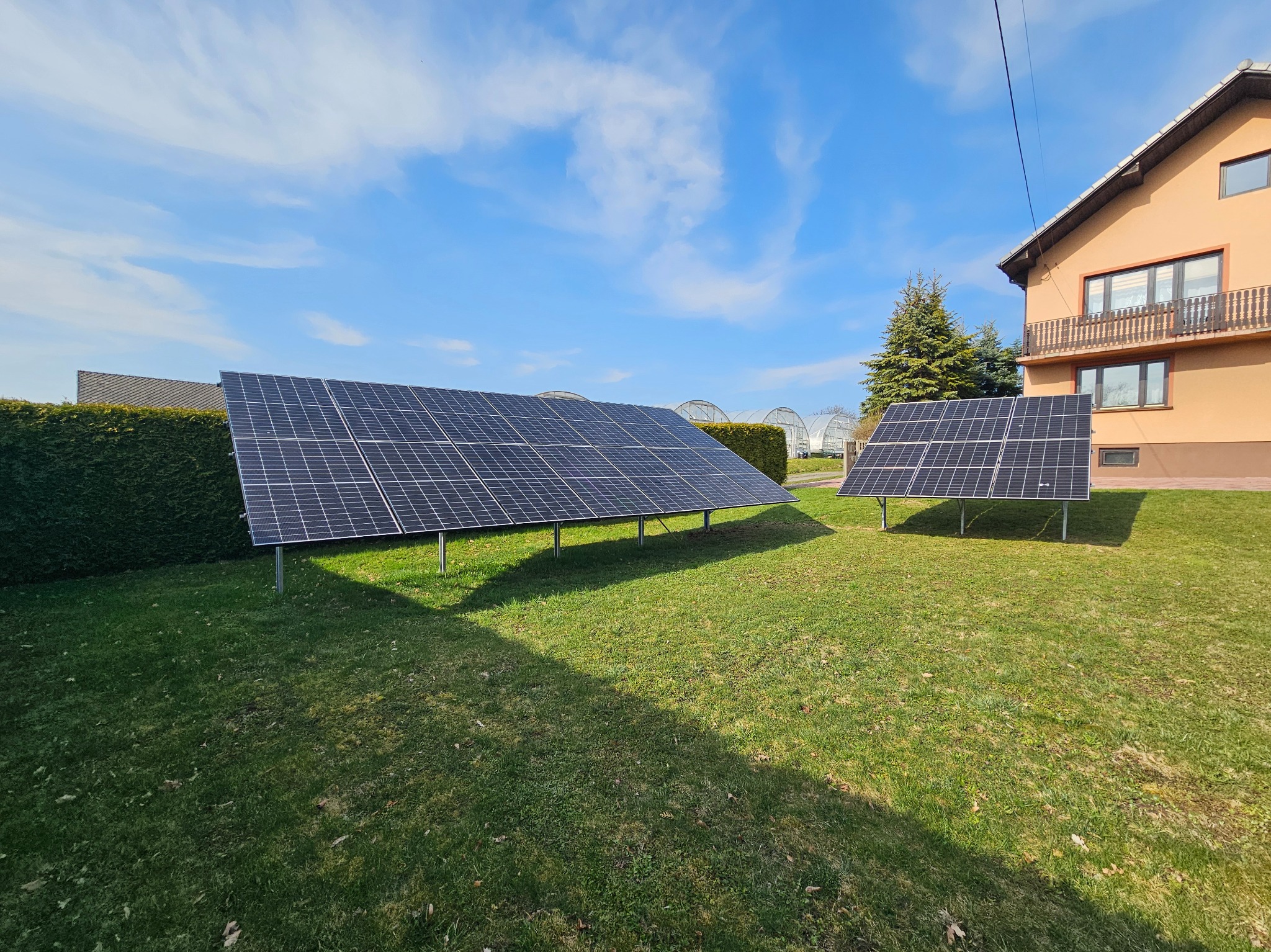 Dwa panele fotowoltaiczne ustawione na trawniku przed domem w słoneczny dzień. W tle widać szklarnie i budynek mieszkalny.