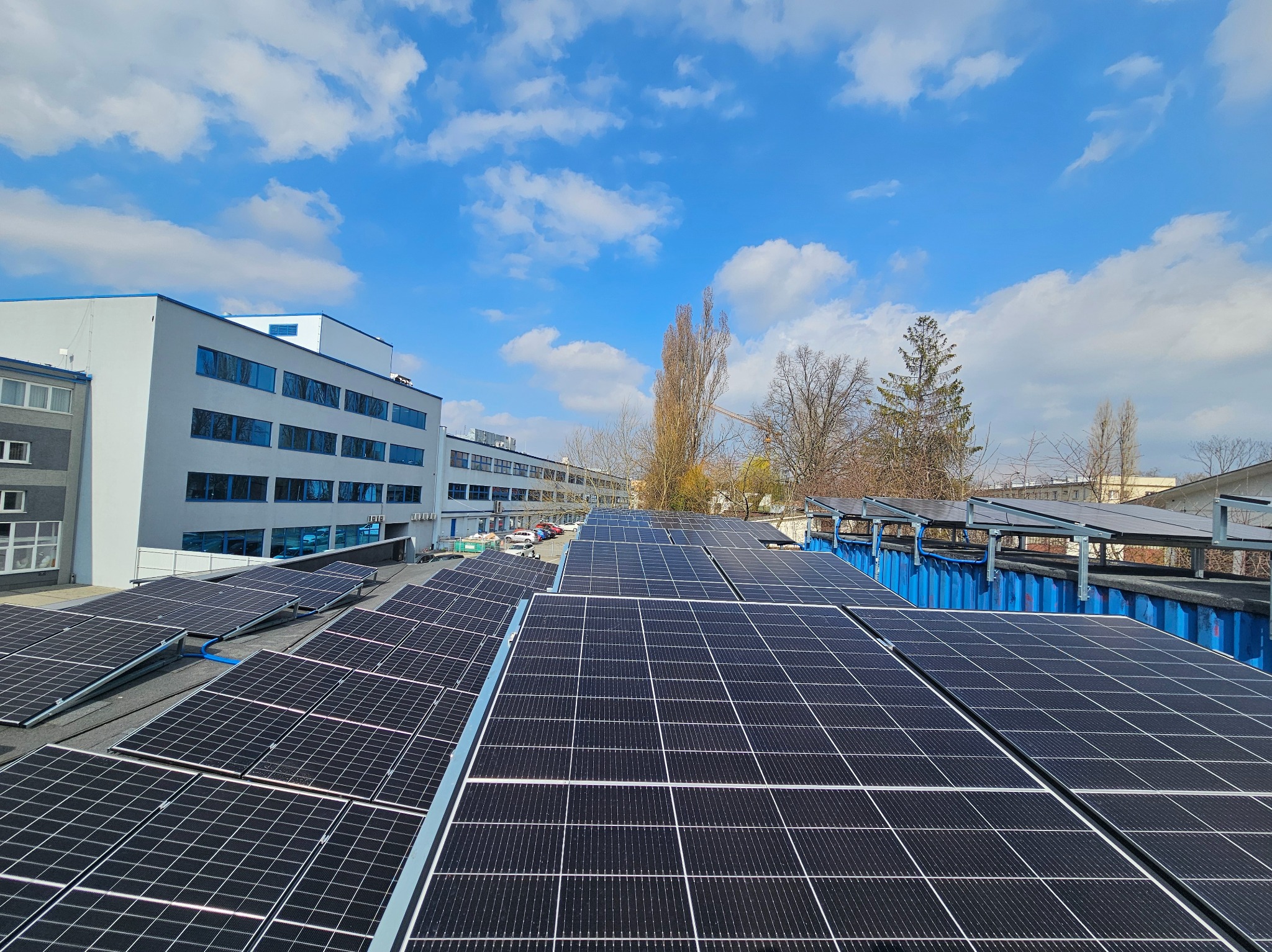 Instalacja paneli słonecznych na dachu budynku przemysłowego. Widok z góry, błękitne niebo z chmurami w tle. Panele równomiernie rozmieszczone, widoczne elementy konstrukcyjne.