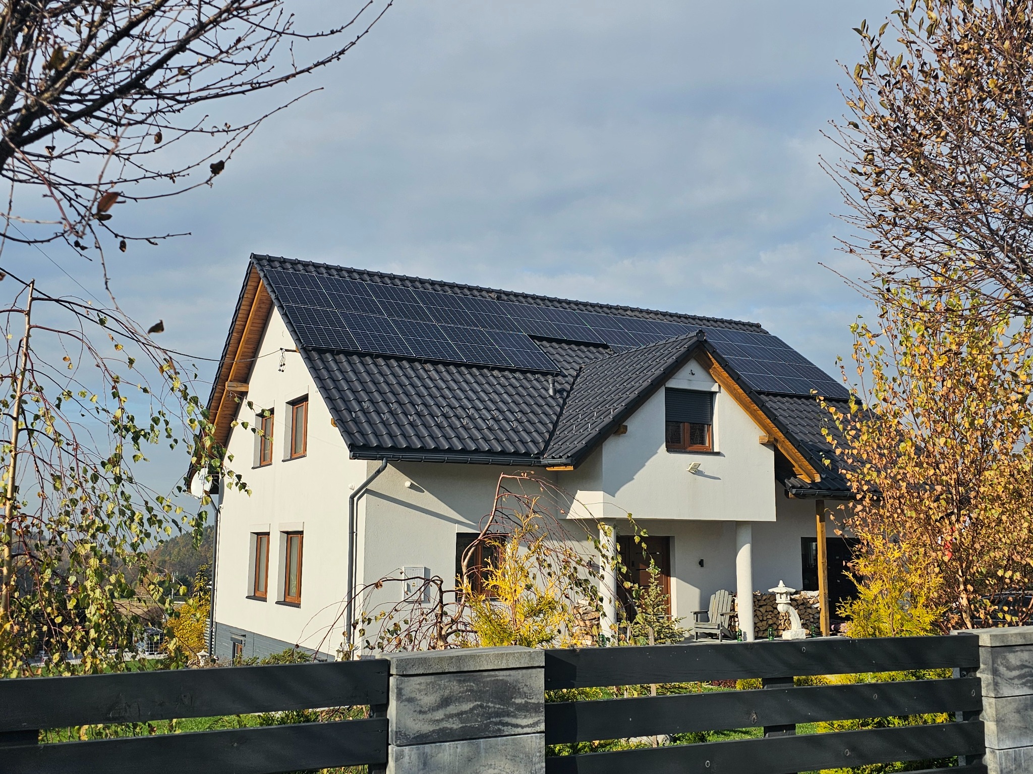 Biały dom jednorodzinny z ciemną dachówką i panelami fotowoltaicznymi na dachu. Widok zza ogrodzenia, jesienne drzewa w tle, pochmurne niebo.