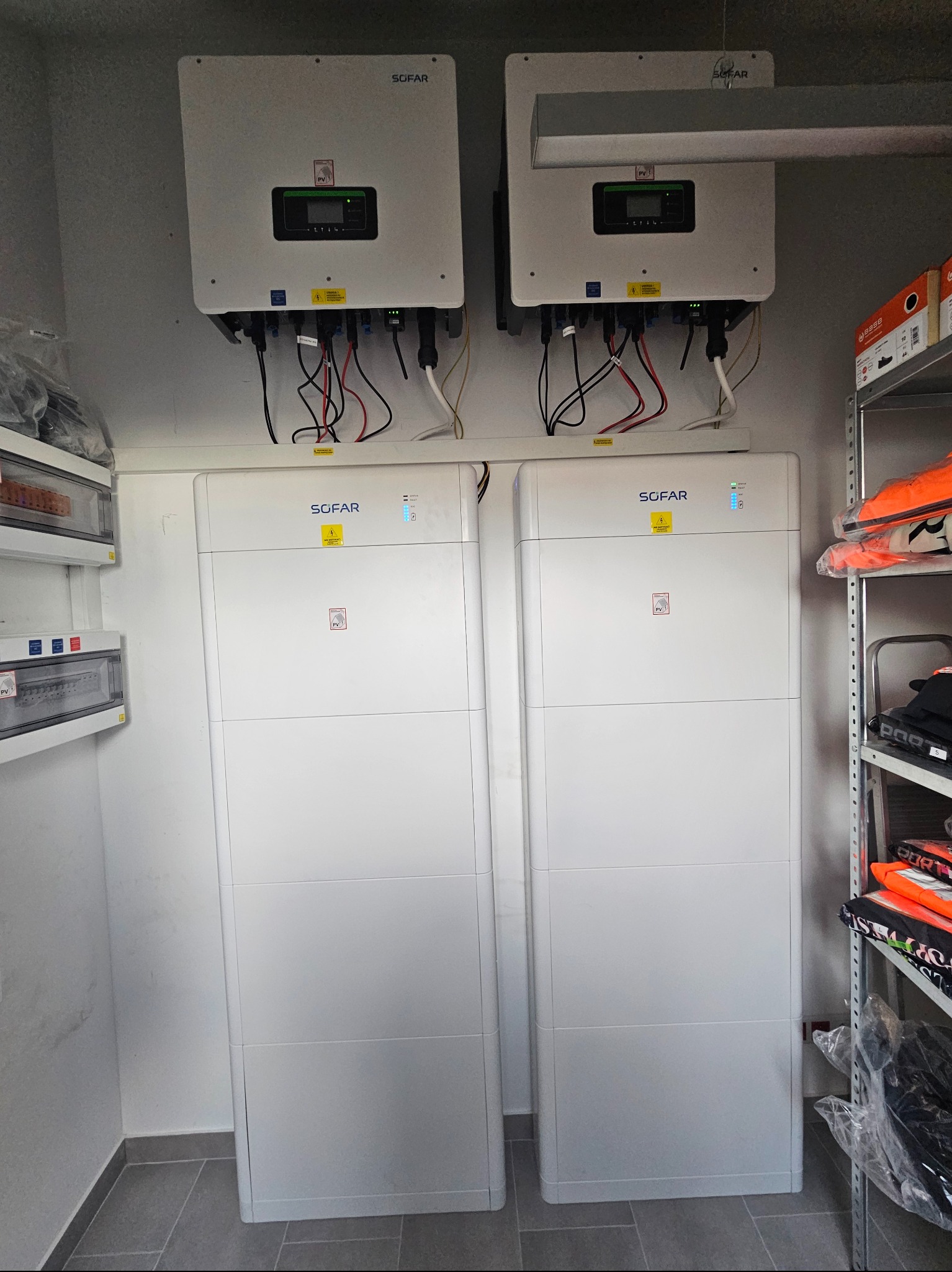 Dwa białe inwertery i dwa magazyny energii SOFAR zamontowane w pomieszczeniu. Widoczne kable i instalacja elektryczna. Półka z ubraniami w tle.