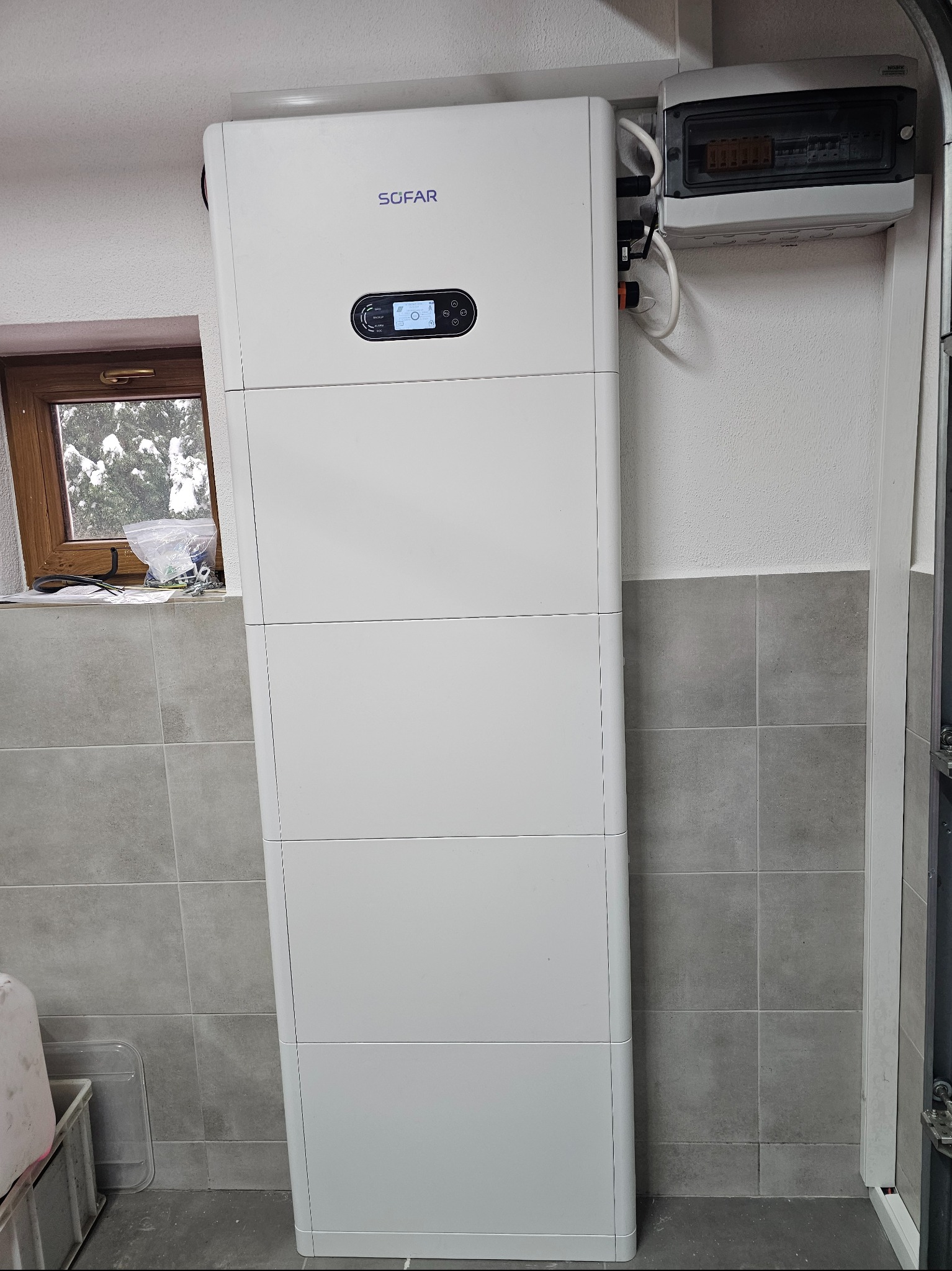 Biały, pionowy magazyn energii SOFAR zamontowany w pomieszczeniu z szarą glazurą. Widoczna skrzynka elektryczna i okno z zimowym krajobrazem.