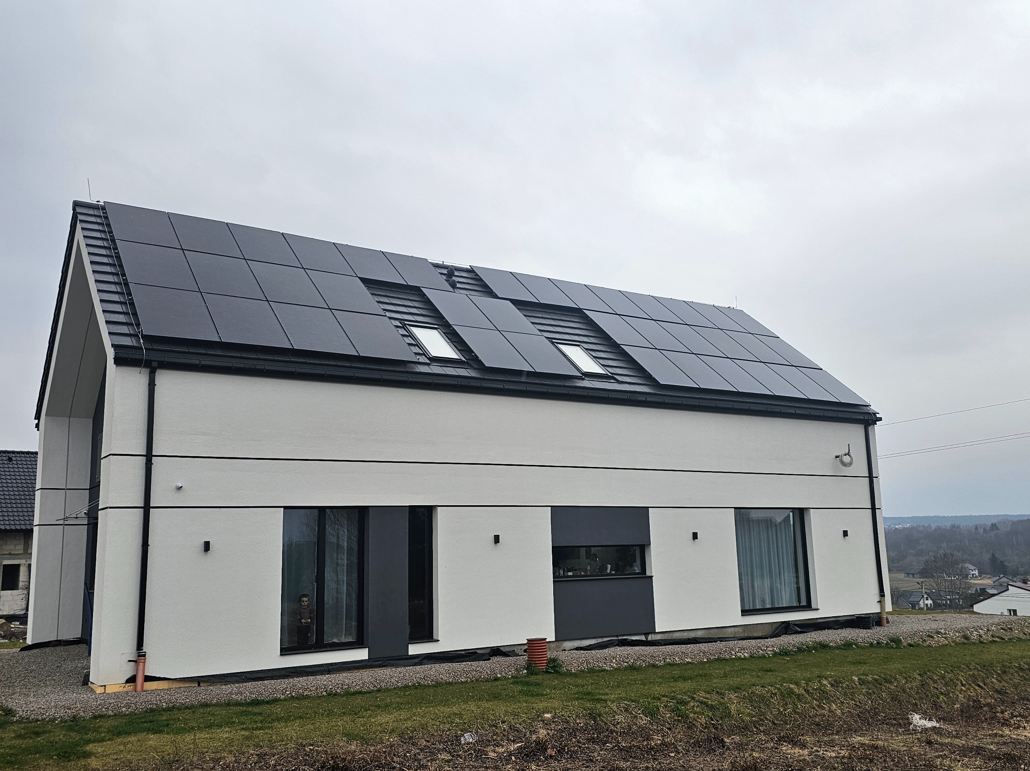 Nowoczesny dom jednorodzinny z panelami fotowoltaicznymi na dachu, widok z zewnątrz w pochmurny dzień. Minimalistyczna architektura, ciemne ramy okienne.