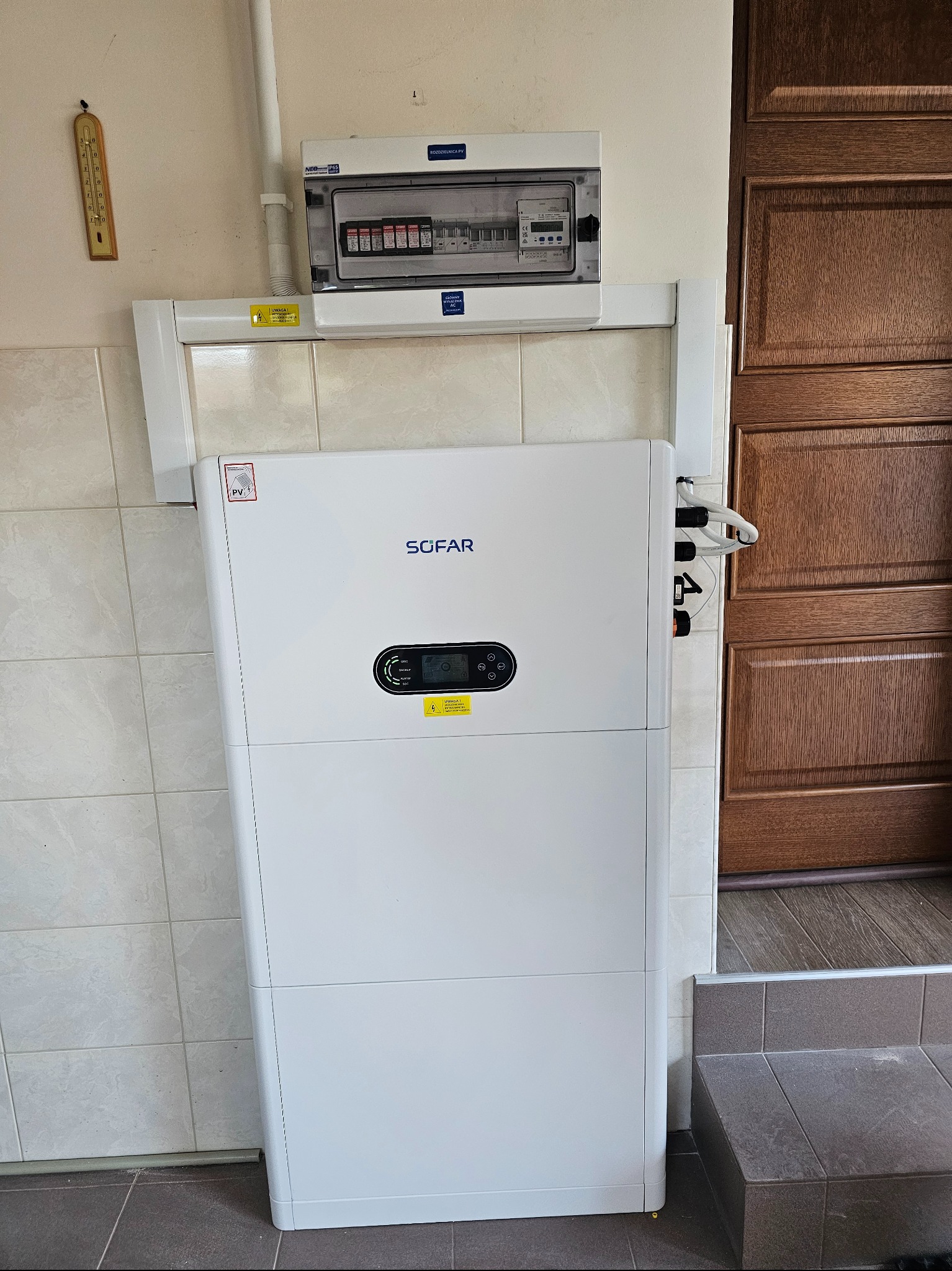 Biały inwerter Sofar zamontowany w pomieszczeniu z brązowymi drzwiami i rozdzielnicą elektryczną. Widoczne kable i elementy instalacji.