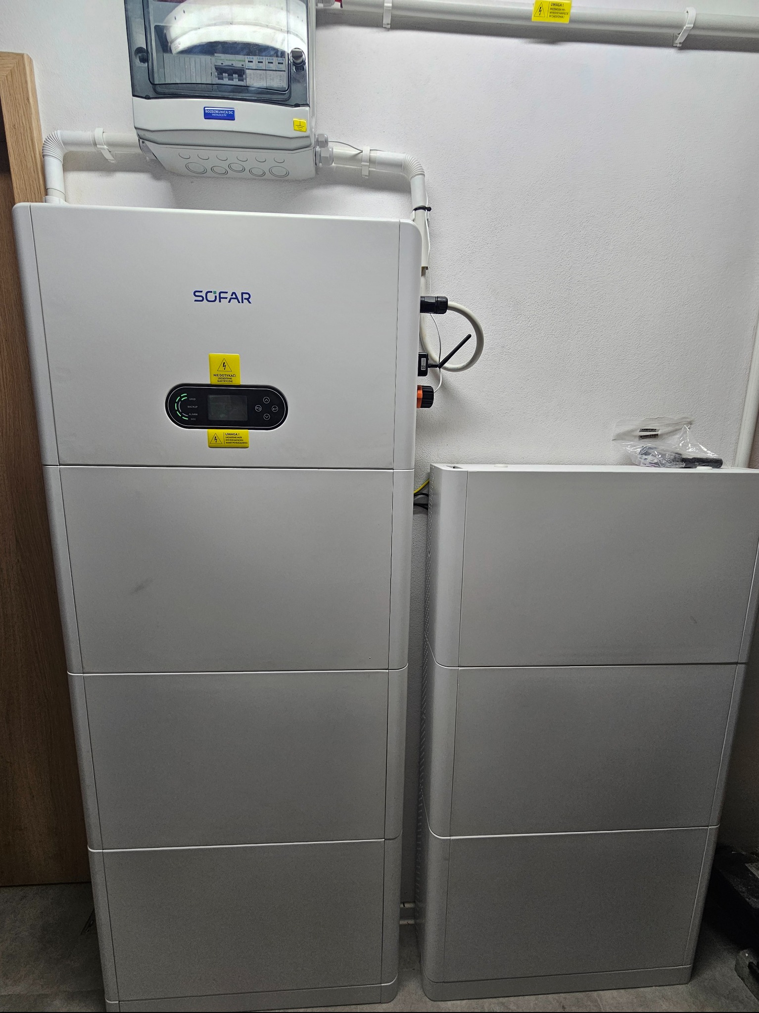 Biały magazyn energii SOFAR z podłączonymi przewodami i skrzynką elektryczną na ścianie. Widoczne naklejki ostrzegawcze i elementy montażowe.