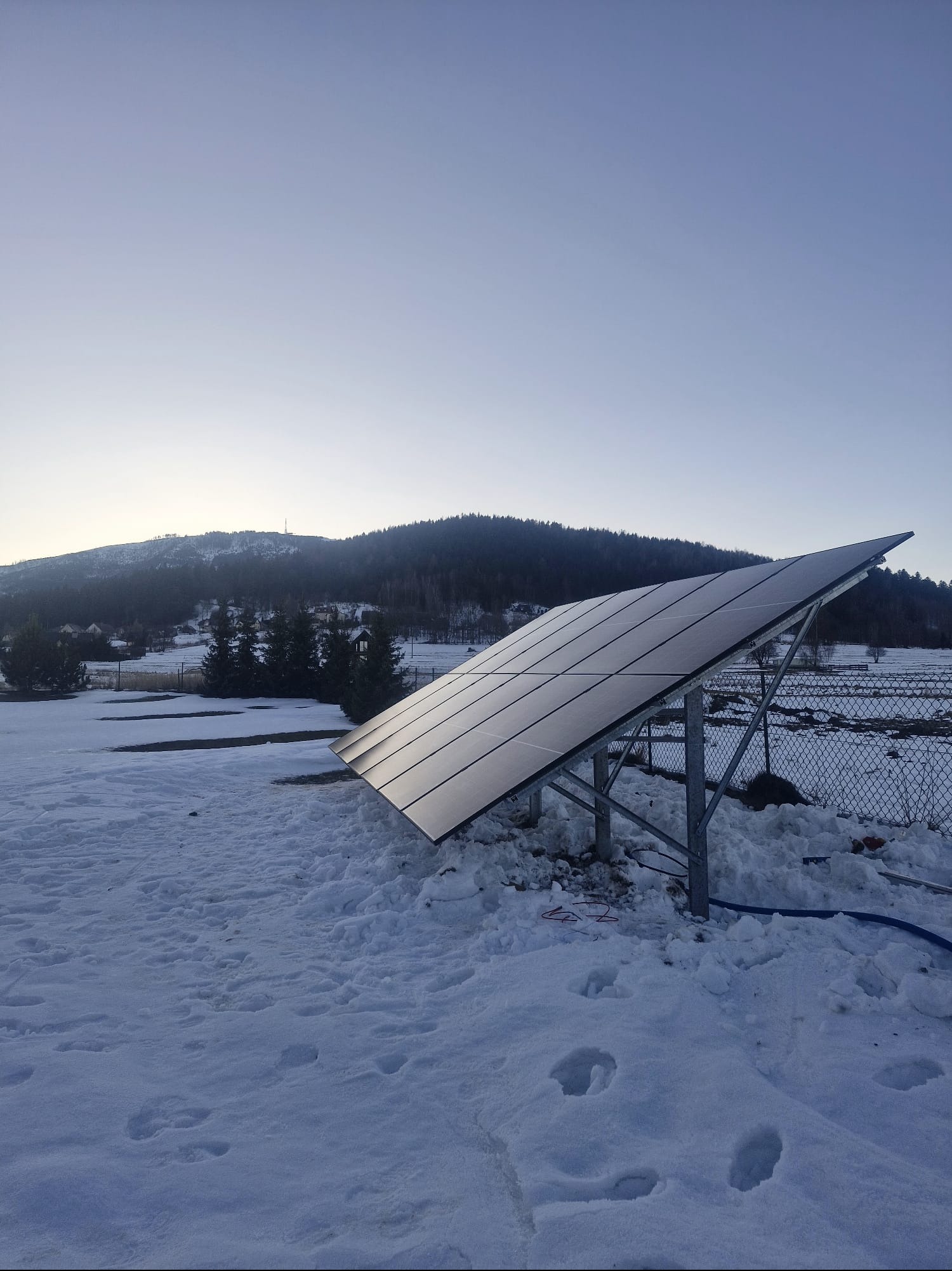 Panel fotowoltaiczny na metalowej konstrukcji, pokryty śniegiem, z widokiem na zalesione wzgórze w tle. Poranek, delikatne światło.