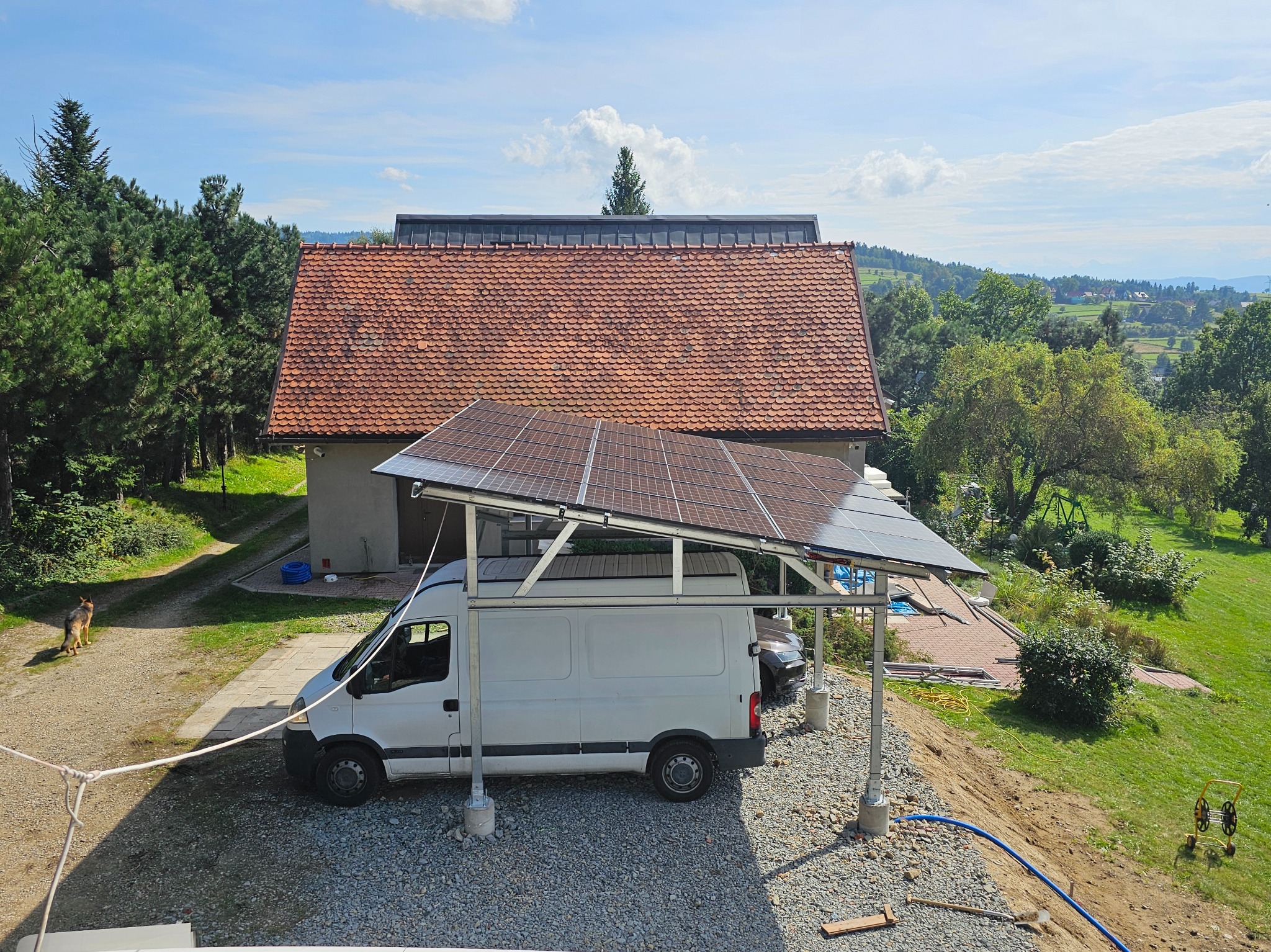 Instalacja fotowoltaiczna nad samochodem dostawczym na tle domu z czerwoną dachówką i zieleni. Nietypowe rozwiązanie z panelami słonecznymi.