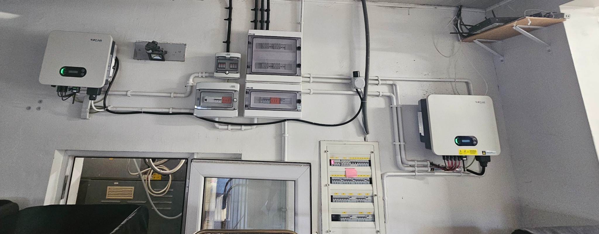 Dwa inwertery SOFAR na ścianie, obok rozdzielnice elektryczne i rury instalacyjne. Widoczne okablowanie i elementy montażowe. Całość w pomieszczeniu o białych ścianach.