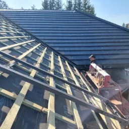 Montaż łat i kontrłat na folii dachowej, widoczny fragment pokrycia dachowego z szarej dachówki ceramicznej oraz pracownik przy maszynie do cięcia dachówek.