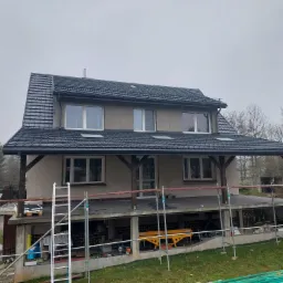 Dom w trakcie wymiany dachu z czarnej dachówki, widoczne rusztowania i sprzęt budowlany na tle pochmurnego nieba.