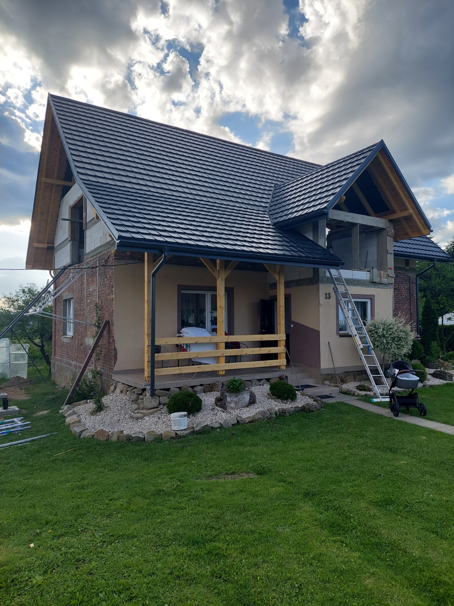 Dom jednorodzinny w trakcie rozbudowy z nowym, ciemnoszarym pokryciem na części dachu, widoczna drabina oparta o ścianę i wózek dziecięcy na trawniku.