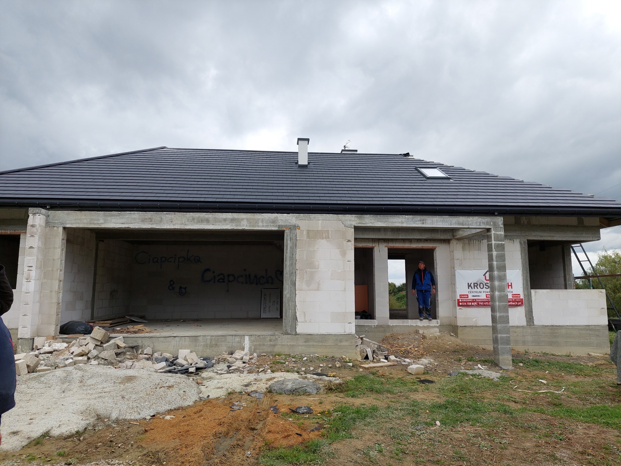 Niedokończony dom jednorodzinny z czarnym dachem z dachówki ceramicznej, widoczne rynny, okno dachowe i komin, ściany z pustaków z napisem i reklamą firmy dekarskiej, teren budowy z gruzem.