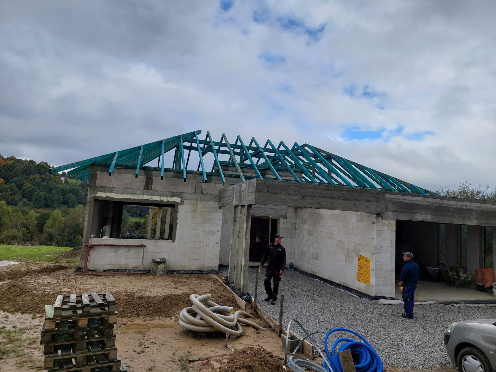 Budowa domu jednorodzinnego w stanie surowym otwartym z widoczną konstrukcją drewnianą dachu pomalowaną na zielono, dwaj pracownicy na placu budowy, palety i zwinięte rury na pierwszym planie...