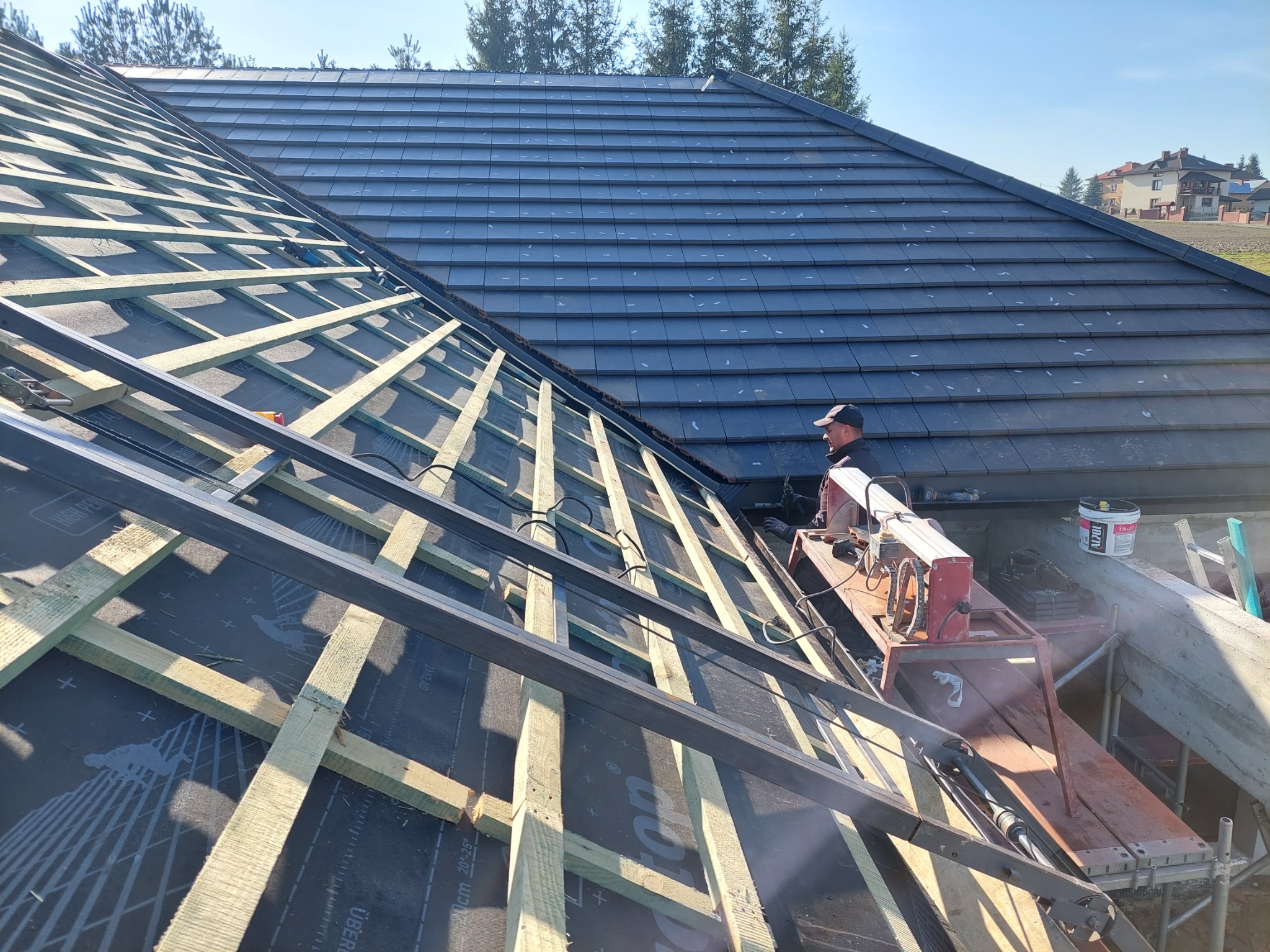 Montaż łat i kontrłat na folii dachowej, widoczny fragment pokrycia dachowego z szarej dachówki ceramicznej oraz pracownik przy maszynie do cięcia dachówek.