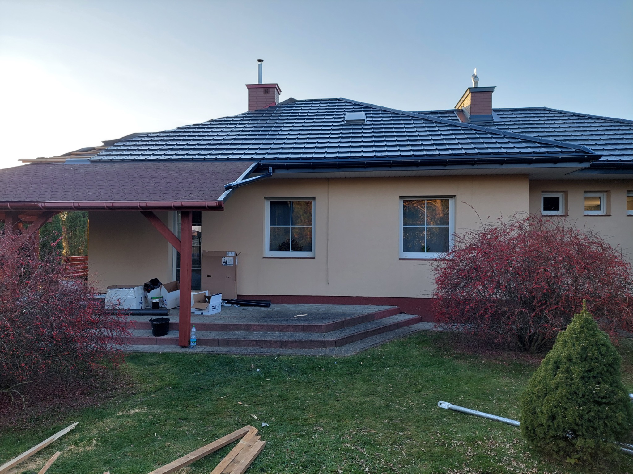 Dom jednorodzinny z dachem pokrytym szarą dachówką bitumiczną, widoczne rynny w kolorze czarnym, fragment drewnianej konstrukcji z boku domu, rozpakowane kartony na tarasie, oraz elementy drewniane...