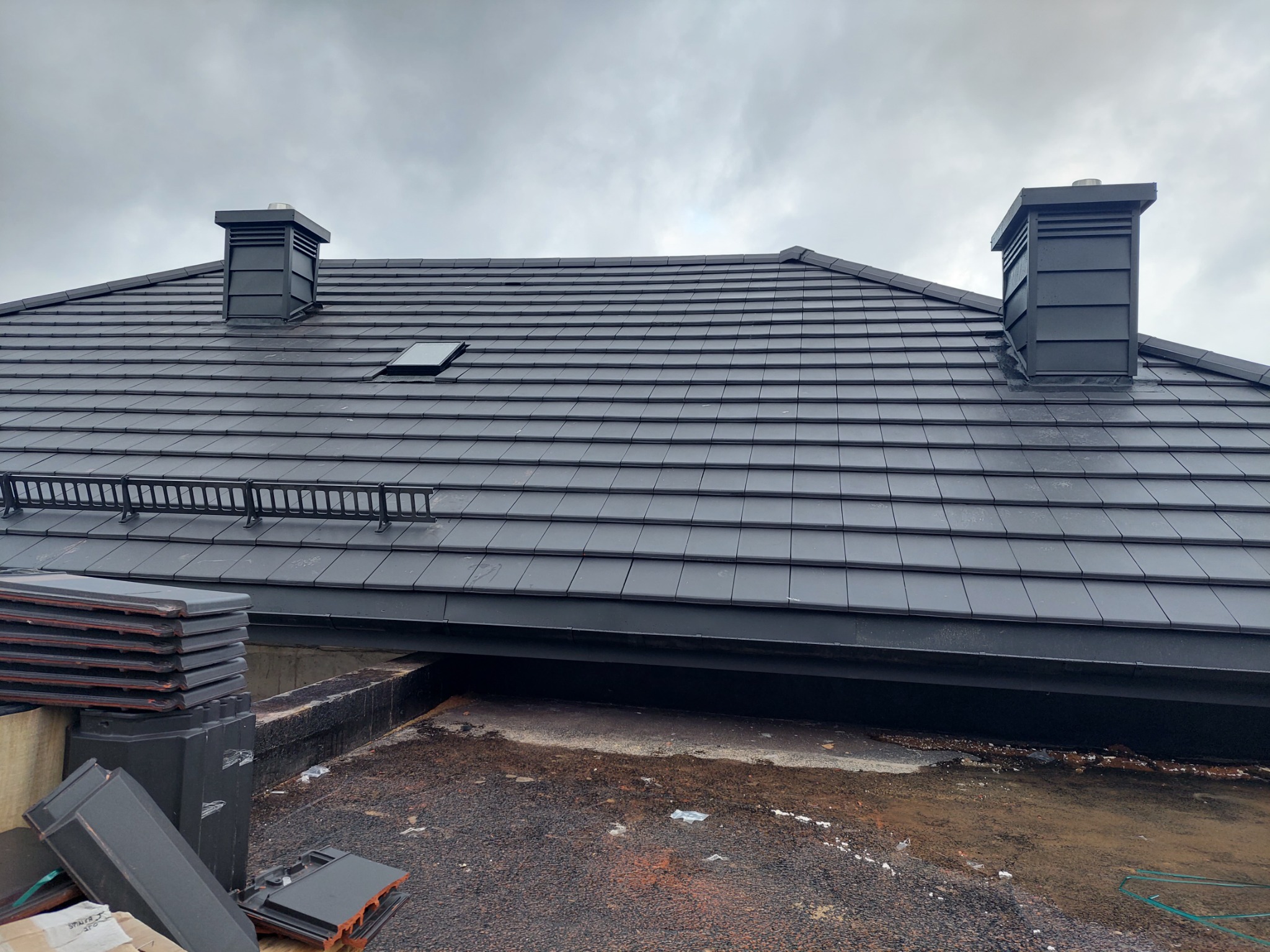 Nowy dach z czarnej dachówki ceramicznej, widoczne dwa kominy wentylacyjne i okno dachowe, na dole stos dachówek przygotowanych do montażu.