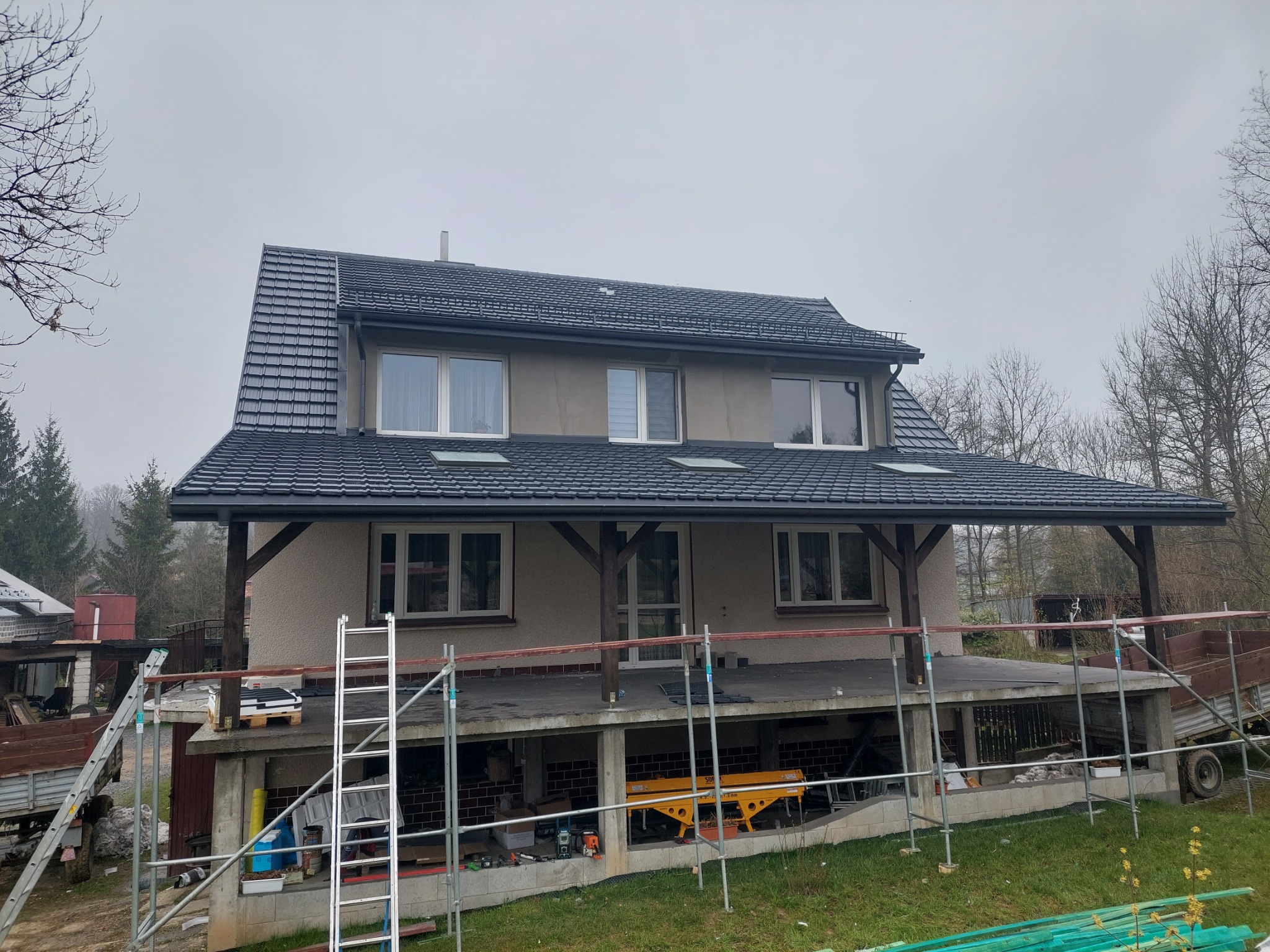 Dom w trakcie wymiany dachu z czarnej dachówki, widoczne rusztowania i sprzęt budowlany na tle pochmurnego nieba.
