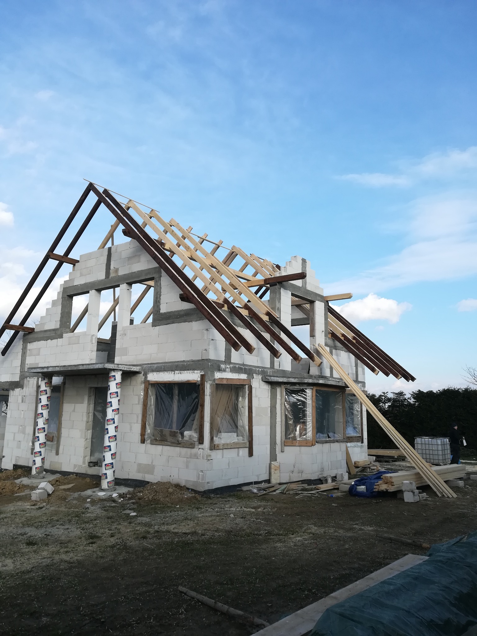 Surowy stan domu jednorodzinnego z bloczków, z widoczną konstrukcją więźby dachowej w trakcie montażu, zabezpieczone okna folią, z drewnem budowlanym na ziemi.