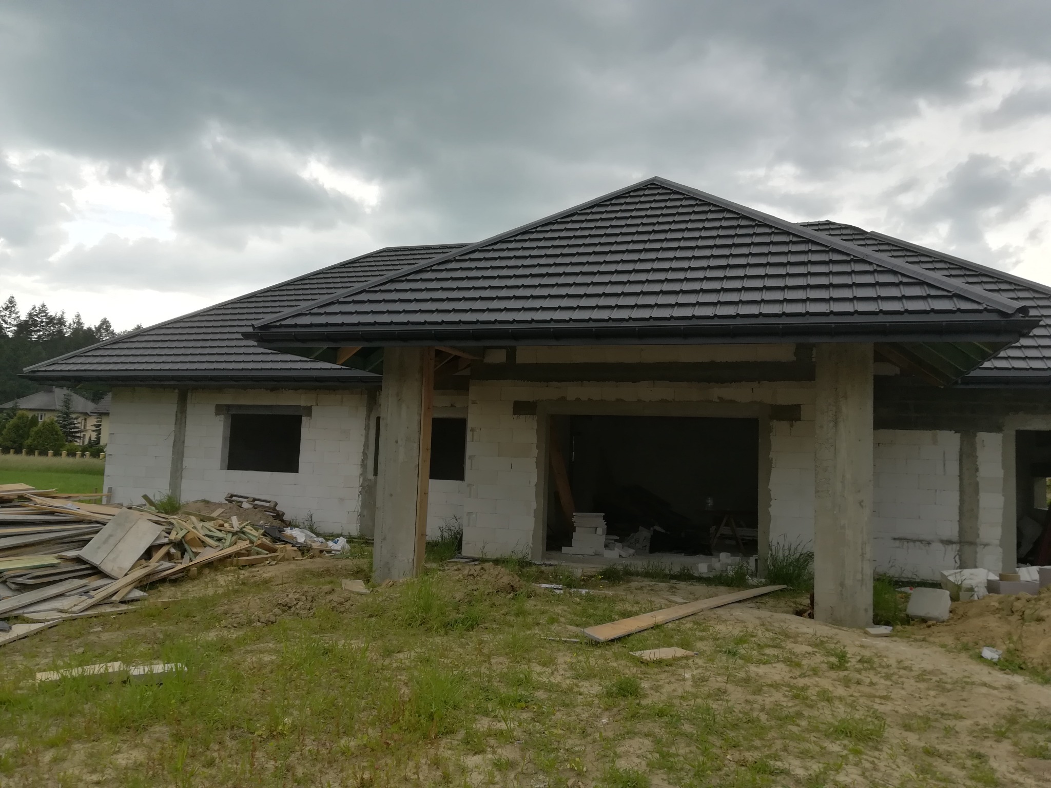 Nowy dom w trakcie budowy z ciemną dachówką i zamontowanymi rynnami, widoczne materiały budowlane i pochmurne niebo.