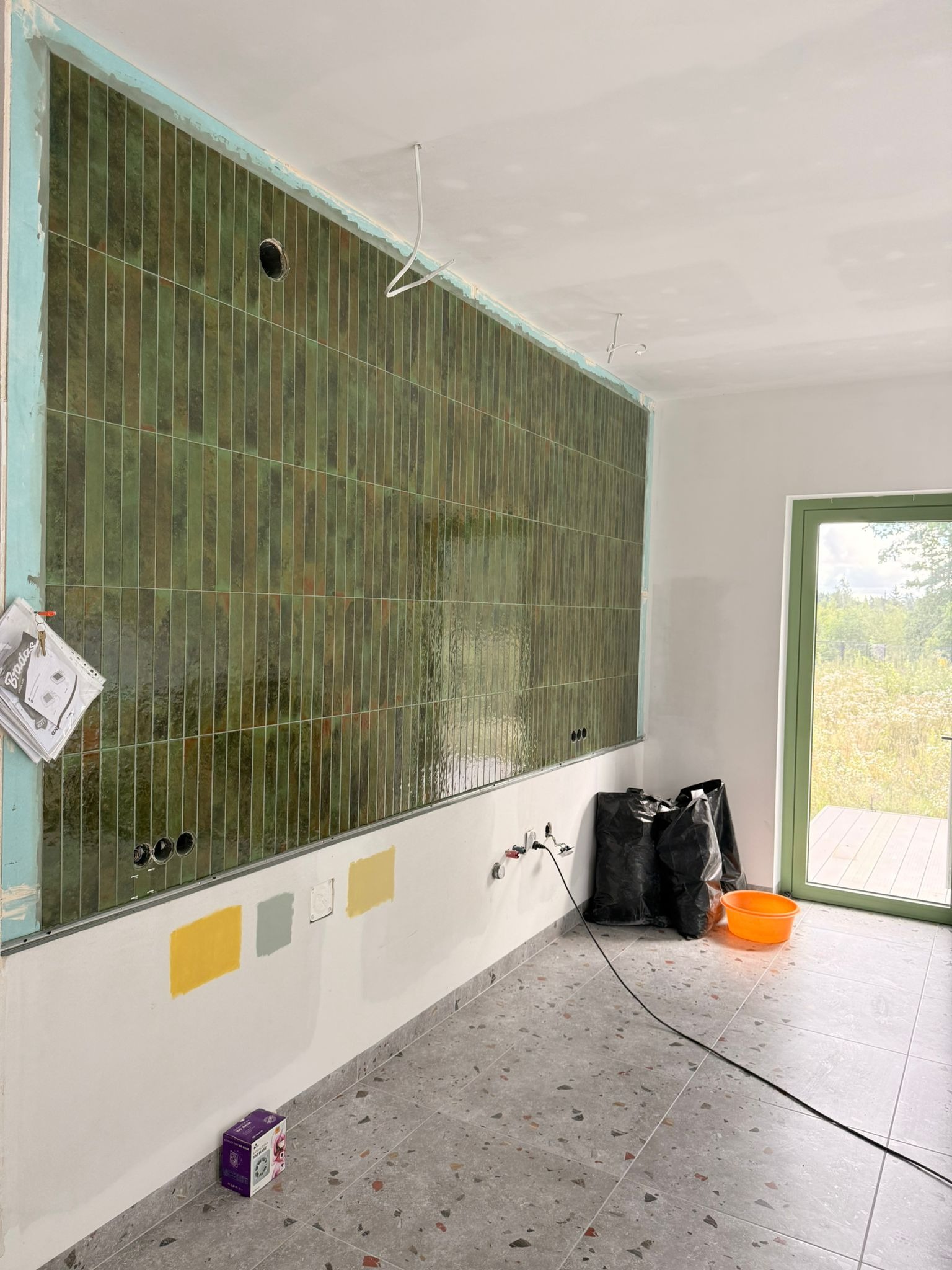 Wnętrze w trakcie wykańczania: zielona, błyszcząca glazura na ścianie, widoczne puszki elektryczne, próbki kolorów farb na ścianie, okno z widokiem na zieleń.