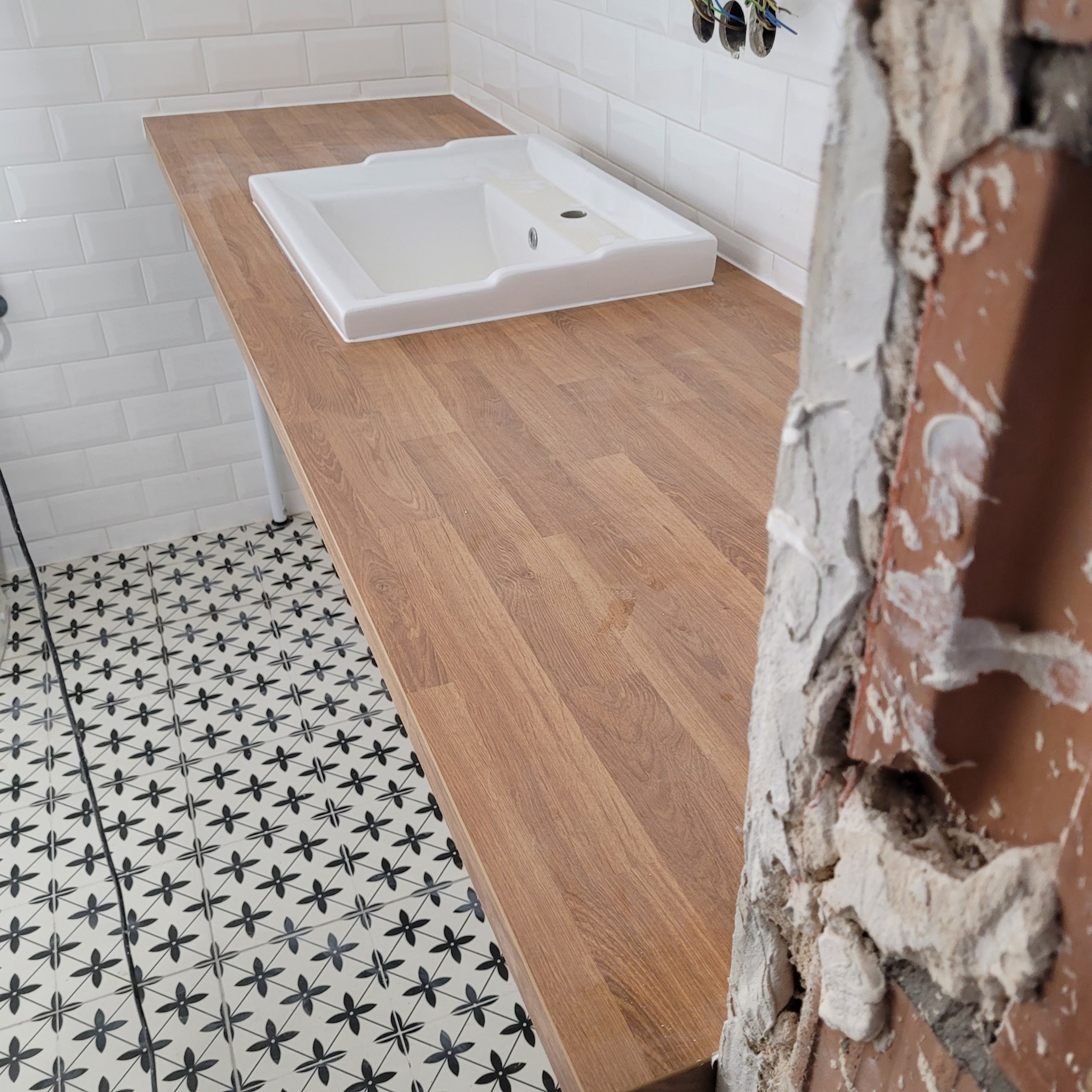 Wykończona drewniana zabudowa umywalki z białą umywalką, białe kafelki na ścianie i wzorzysta podłoga. Fragment ściany w trakcie prac wykończeniowych.