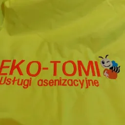 Żółta tkanina z wyhaftowanym logo firmy 'EKO-TOMI Usługi asenizacyjne', przedstawiającym uśmiechniętą pszczołę z dzbankiem miodu.