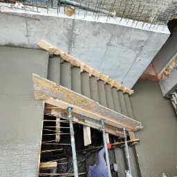 Wylewanie betonowych schodów z drewnianym szalunkiem i metalowymi podporami teleskopowymi, widoczne zbrojenie i fragment pracownika w tle.