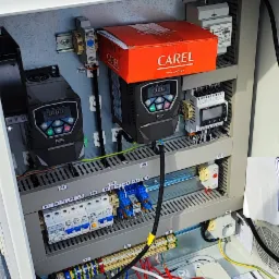 Otwarta szafa sterownicza z dwoma falownikami ELRA, sterownikiem CAREL w czerwonym pudełku, bezpiecznikami, licznymi przewodami i puszką napoju energetycznego Level Up Zero na górze, zainstalowana...