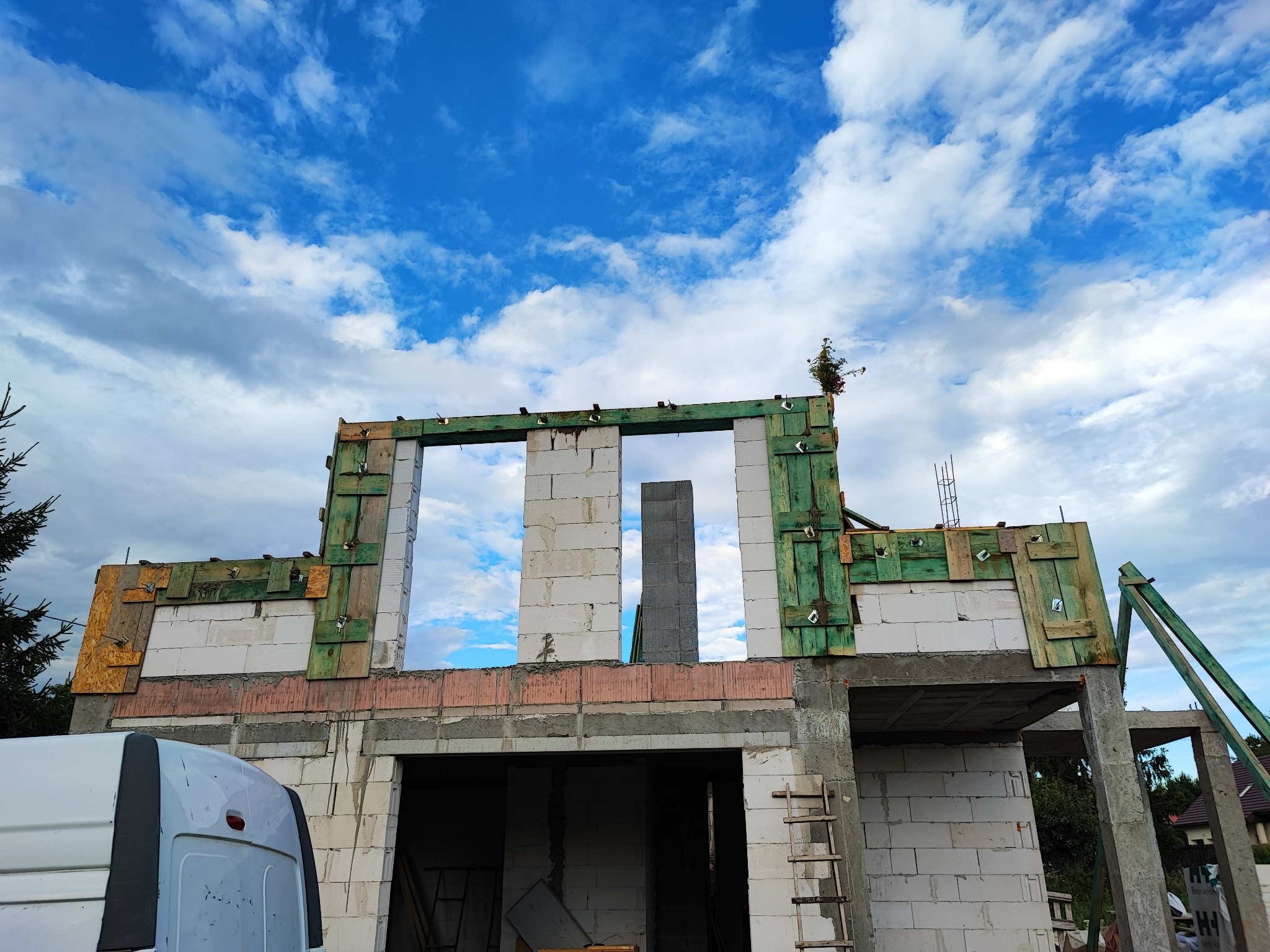 Wznoszenie ścian budynku z betonu komórkowego, widoczne szalunki i podpory, na górze konstrukcji rosnąca roślina, na pierwszym planie fragment białego samochodu dostawczego.