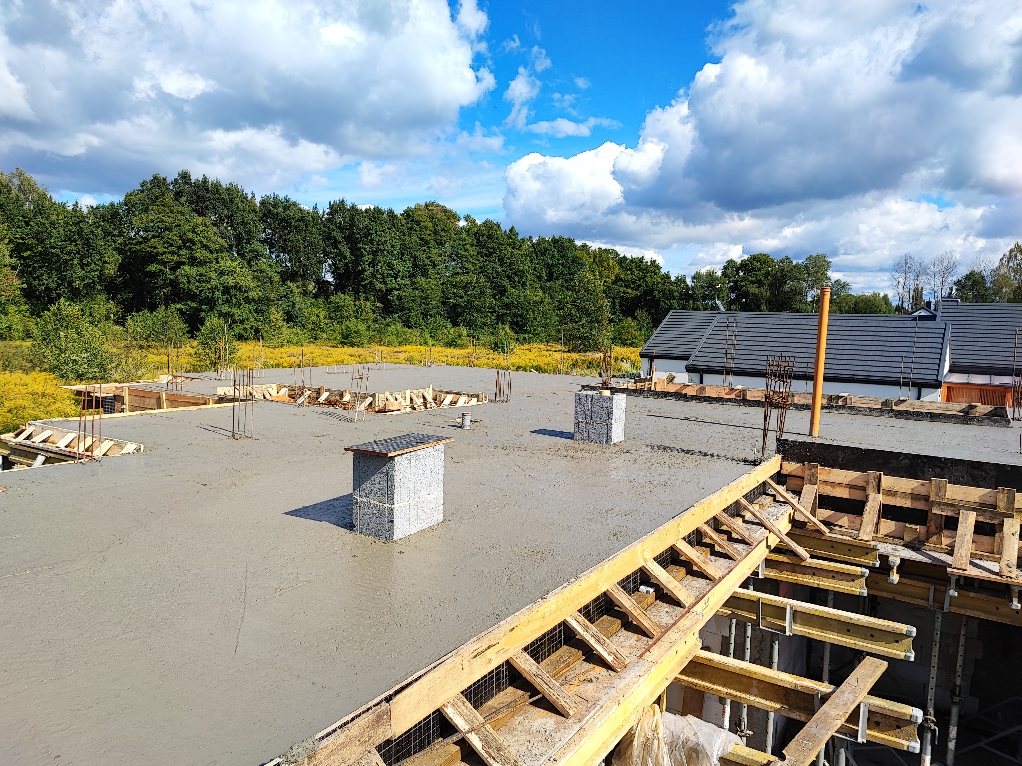 Wylany beton na stropie budynku w trakcie budowy, widoczne szalunki i zbrojenia, w tle budynki i drzewa pod błękitnym niebem z chmurami.