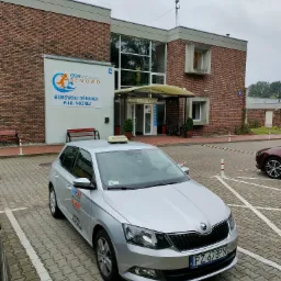 Srebrny samochód z napisem 'Ochrona' na dachu, zaparkowany na parkingu przed budynkiem z cegły z logo Bemowskiego Ośrodka Piłki Nożnej i napisem 'HAPRO Agencja Ochrony', widoczny numer...