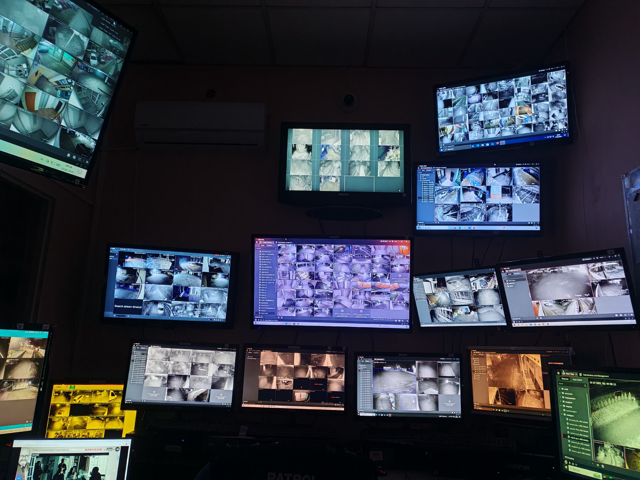 Ściana monitorów w centrum nadzoru wizyjnego, wyświetlających obrazy z wielu kamer CCTV, z widocznym napisem 'PATROL' na biurku operatorskim.