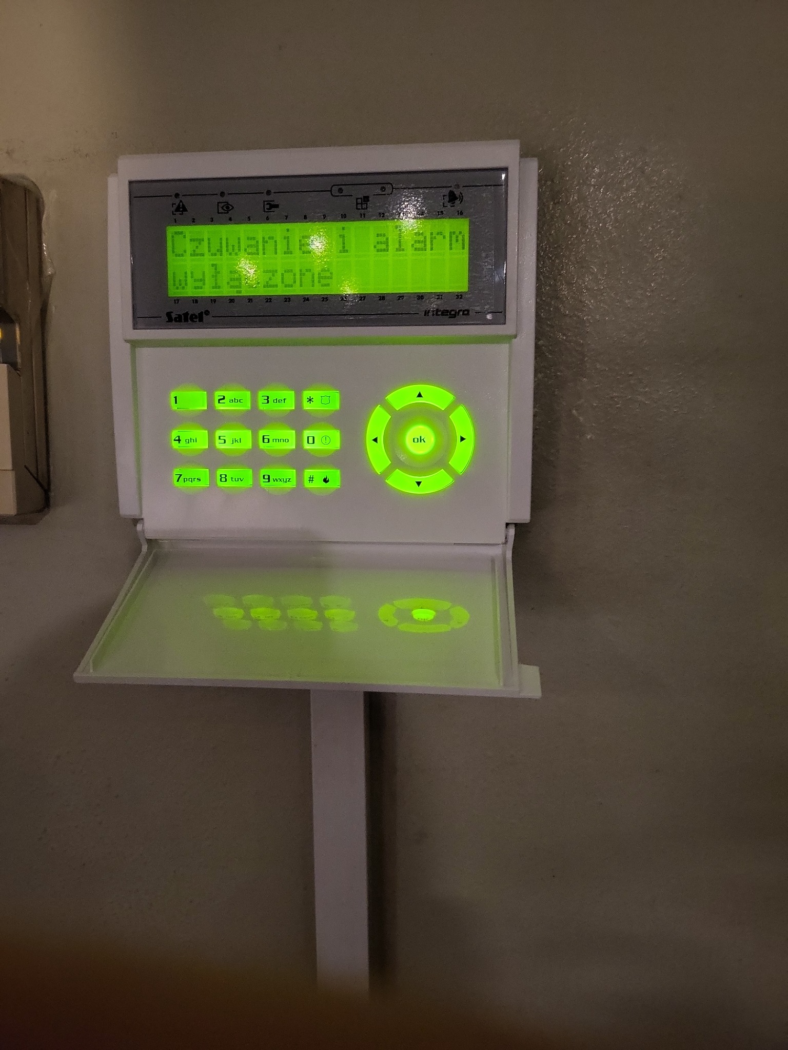 Panel sterowania alarmem z podświetlanymi na zielono przyciskami i wyświetlaczem pokazującym status 'Czuwanie i alarm wyłączone', zamontowany na ścianie o nierównej fakturze.