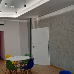 MM Perfekt Maciej Majewski - Wykończone wnętrze biura z dekoracyjną ścianą o fakturze betonu, podświetloną pionowymi paskami LED, białe drzwi i okrągły stół z kolorowymi krzesłami.