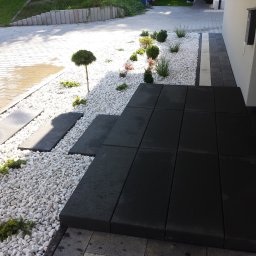 MM Perfekt Maciej Majewski - Nowoczesne wejście do domu z czarnych płyt tarasowych, otoczone białymi kamieniami i ozdobnymi roślinami, prowadzące do drzwi wejściowych.