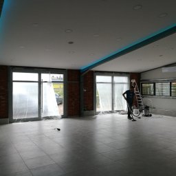 MM Perfekt Maciej Majewski - Przestronne wnętrze z szarą podłogą z płytek, osłoniętymi folią oknami i pracownikiem sprzątającym odkurzaczem przemysłowym, widoczne elementy ceglanej ściany i oświetlenie LED na suficie.