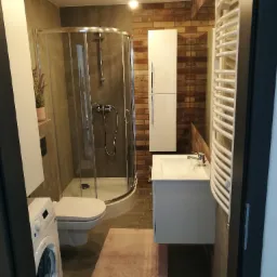 Nowoczesna łazienka z kabiną prysznicową, wiszącą szafką z umywalką, grzejnikiem drabinkowym, pralką i dekoracyjną ścianą z cegły.