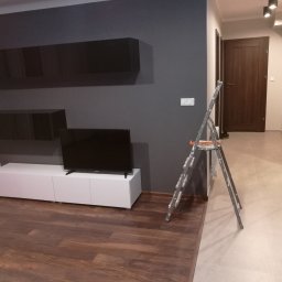 MM Perfekt Maciej Majewski - Pokój po świeżym malowaniu ścian na ciemny grafit, widoczna drabina malarska, biała szafka RTV pod telewizorem, drewniana podłoga, drzwi w tle.
