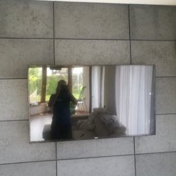 MM Perfekt Maciej Majewski - Szara ściana z dekoracyjnych paneli imitujących beton, z zawieszonym telewizorem odbijającym wnętrze pokoju i osobę fotografującą, klimatyzator zamontowany na górze po prawej stronie.