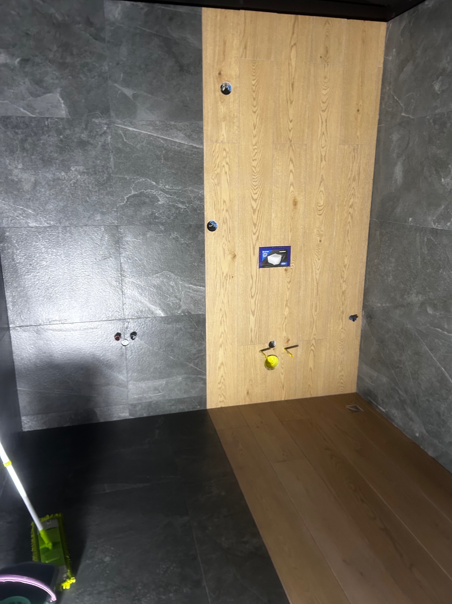 Wnętrze łazienki w trakcie prac wykończeniowych: ściana z szarych płytek imitujących kamień, drewniana zabudowa stelaża podtynkowego WC z widocznymi otworami na instalacje i naklejoną instrukcją...