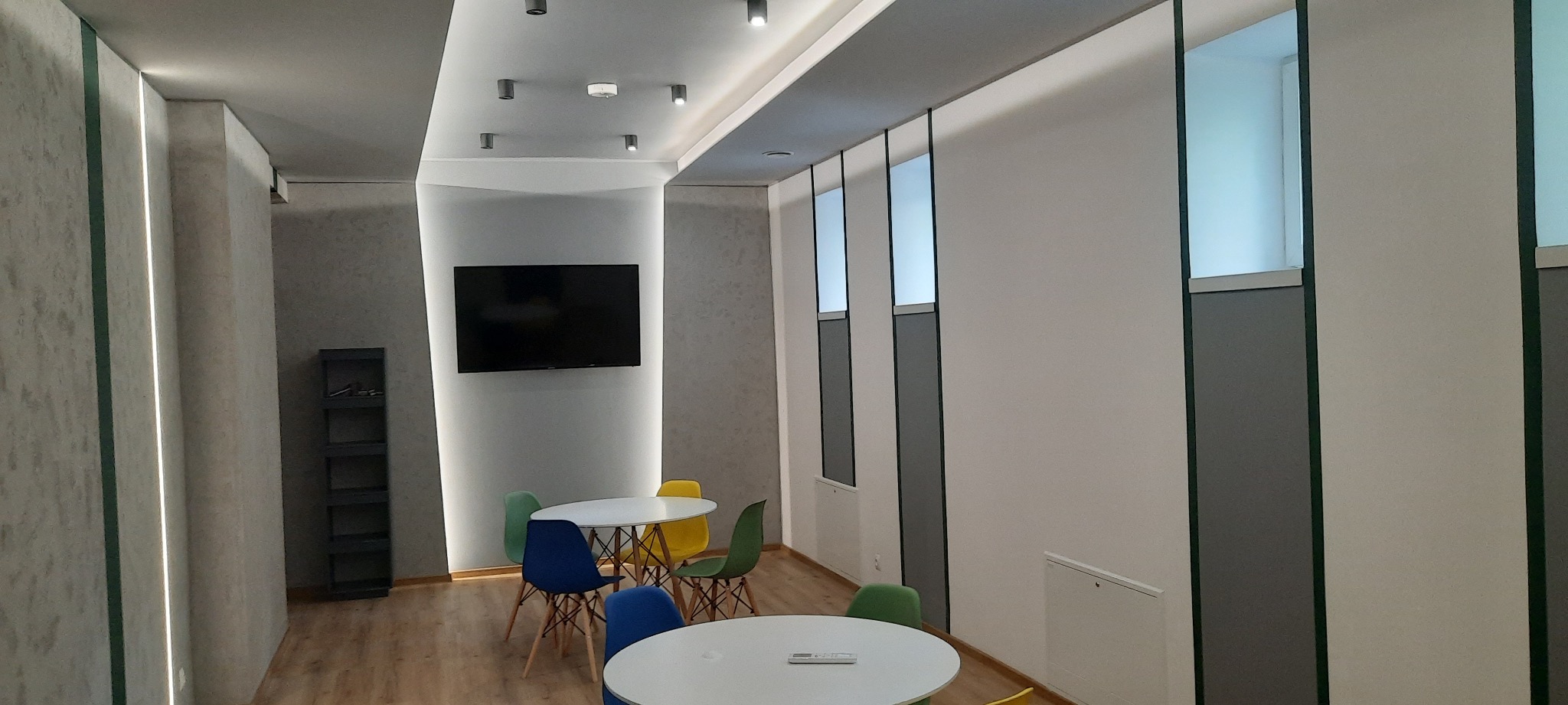 Nowoczesna przestrzeń biurowa z dwoma okrągłymi stołami otoczonymi kolorowymi krzesłami, minimalistyczne oświetlenie sufitowe, ściany w odcieniach szarości i bieli z pionowymi panelami i wnękami...